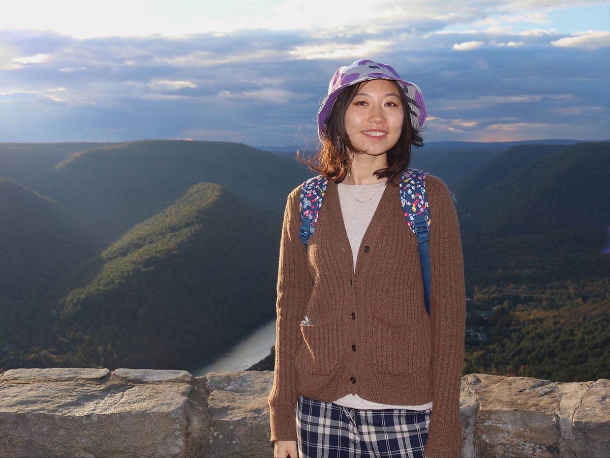 Shuyu Chang standing before a stone wall, with a river and mountains in the background